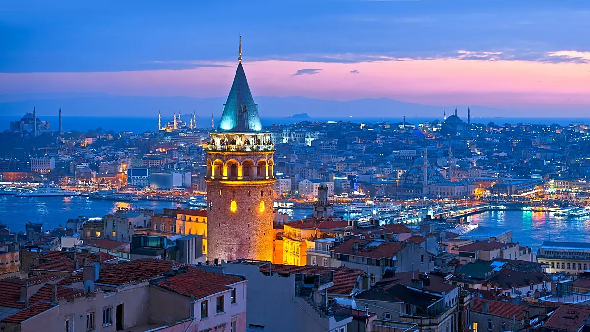 Galata Tower glowing over Istanbul's rooftops at blue hour, with the Golden Horn and old city lights behind